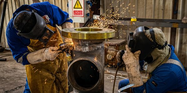 Two welders in protective gear working on metal pipe with sparks flying.