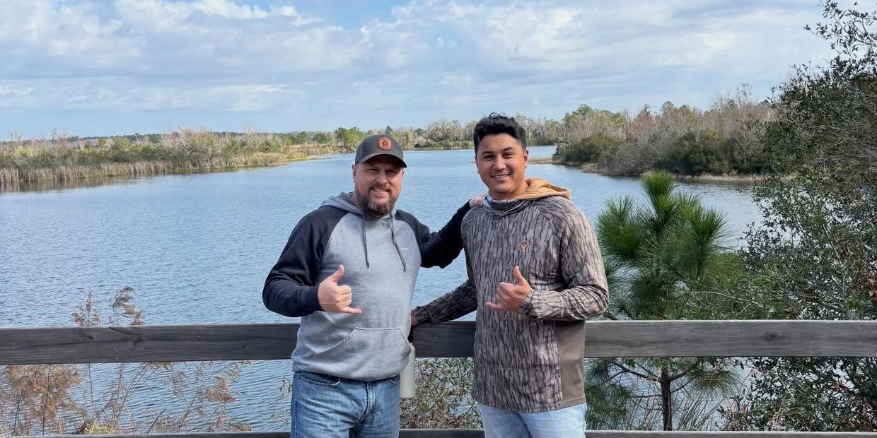 Two men smiling and posing by a lake with a wooden railing and cloudy sky.