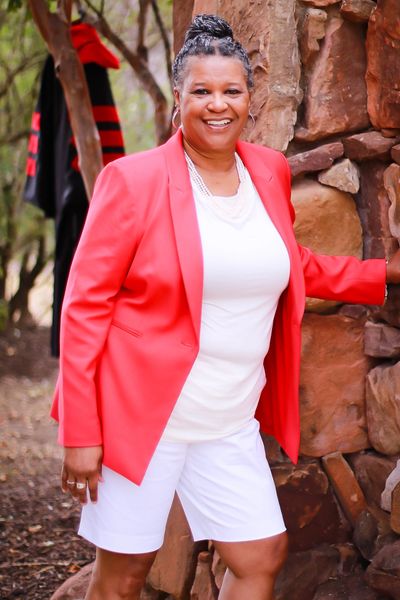 Woman in a bright coral blazer and white outfit smiling outdoors by a stone wall.