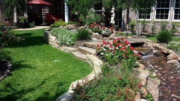A lush garden with stone steps, colorful flowers, and a small water feature near a brick house.