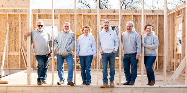 Group of six people posing in a wooden house frame under construction.