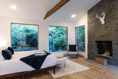 Cozy bedroom with large windows overlooking lush greenery and a stone fireplace.