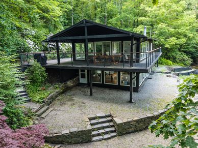 Modern two-story house with wraparound deck surrounded by lush greenery.