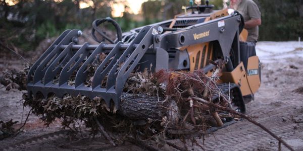 A man operates a Vermeer machine clearing branches and logs outdoors.