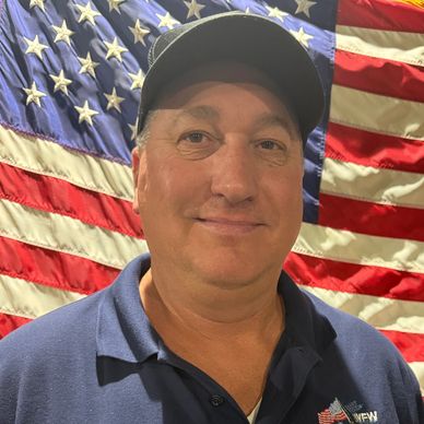 Man smiling in front of an American flag wearing a VFW shirt.