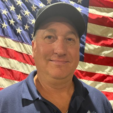 Man smiling in front of an American flag wearing a VFW shirt.