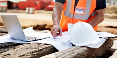 Engineer reviewing construction plans with laptop and helmet on site.