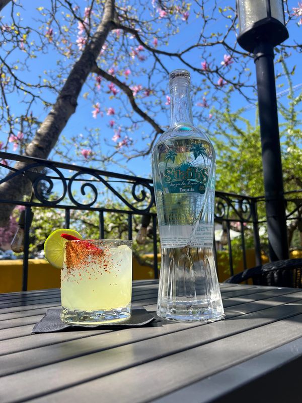A tequila bottle and a spicy cocktail on an outdoor table with a bright blue sky.