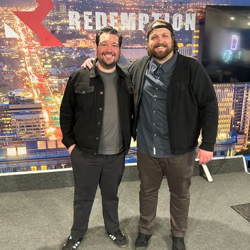 Two smiling men posing in front of a cityscape backdrop with the word 'Redemption'.