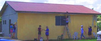People painting the exterior of a yellow house with a red roof.