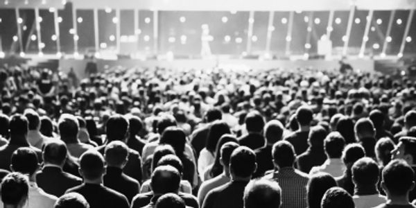 Black and white photo of a large audience facing a stage with bright lights.