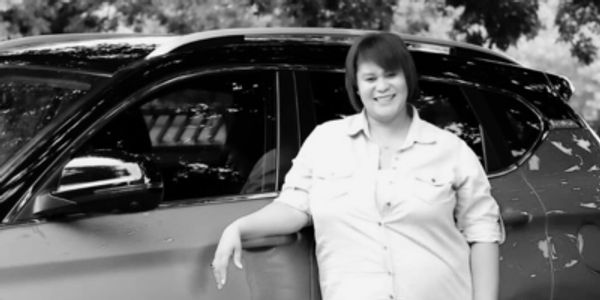 A smiling woman leans against a car in an outdoor setting.