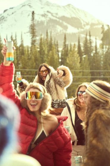 Friends partying outdoors in winter gear with snowy mountains behind.