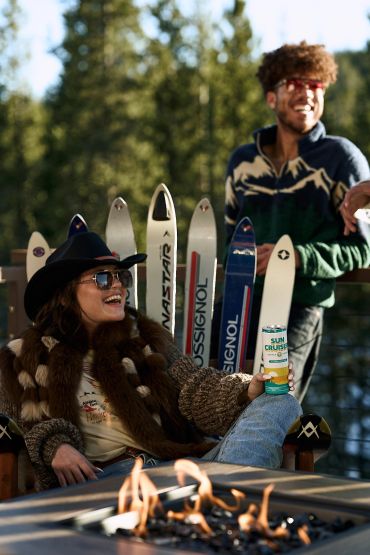 Happy woman in fur jacket enjoying drink near firepit with skis and man.