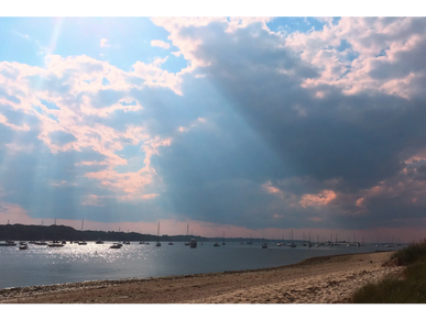 Sun rays break through clouds over a calm beach with boats on the water.