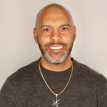 Smiling bald man with a beard wearing a dark sweater and a gold necklace with a scissors pendant.