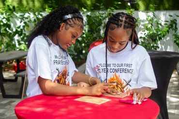 Two girls in event shirts engaged in an activity at a red table outdoors.
