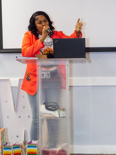 Woman in orange blazer speaking at a podium with a laptop.