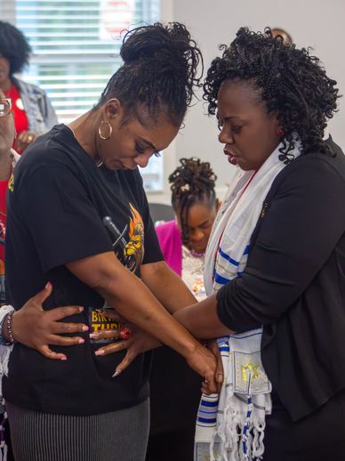 Two women holding hands in a moment of prayer or support indoors.