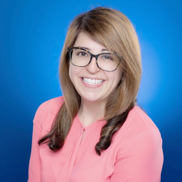 Smiling woman with glasses and curled hair in a pink blouse against a blue background.