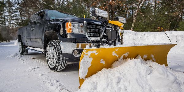Pickup truck with a yellow snow plow clearing a snow-covered driveway surrounded by trees.