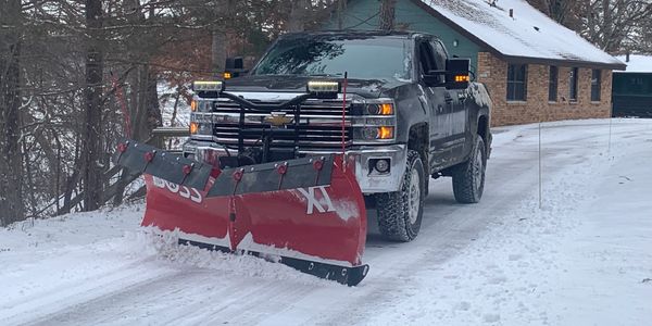 Snow plow truck clearing a residential driveway near a brick home in a wooded, snowy area.