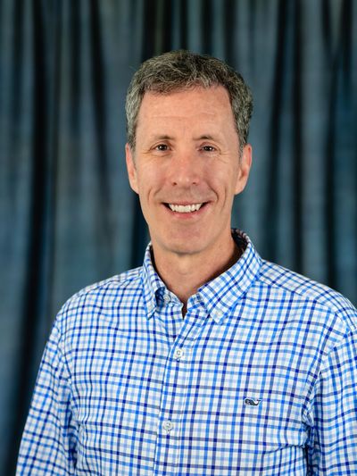 Smiling man in a blue checkered shirt posed against a dark curtain background.