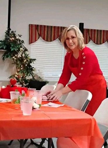 A smiling woman in a red dress sets a table decorated for a festive occasion.