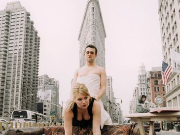 Man wrapped in white stands on a woman leaning over a couch outdoors in a city.