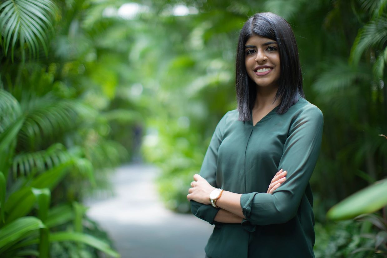 Confident woman in green shirt smiling outdoors with lush greenery.