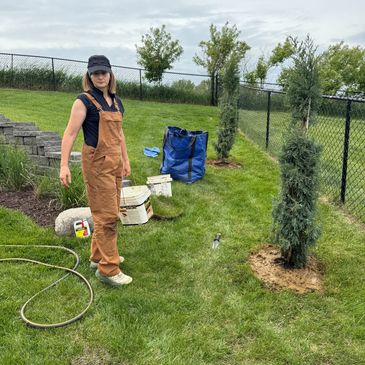 Person in brown overalls planting trees in a grassy yard.