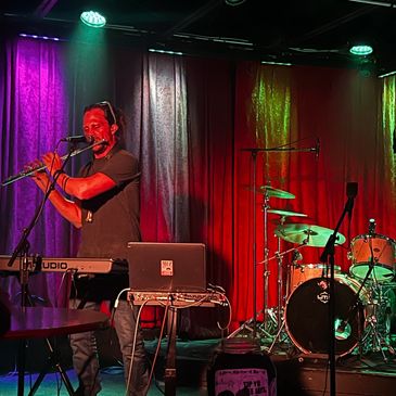 Musician playing flute on stage with colorful lights and drum set in background.