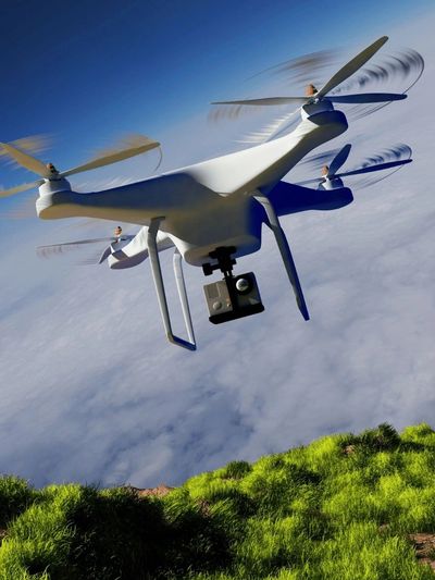 A white quadcopter drone with a camera flying over green terrain under a blue sky.