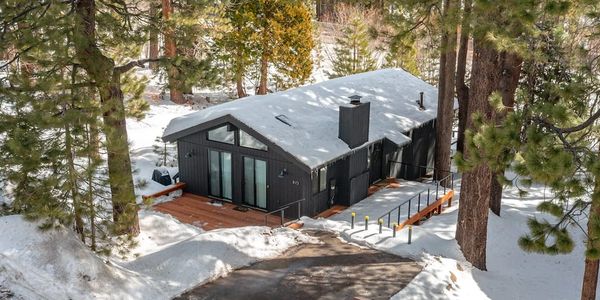 Modern black cabin with snow-covered roof surrounded by pine trees.