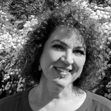 Curly-haired woman smiling outdoors with flowers in the background.
