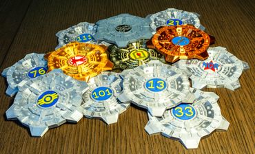 Colorful Fallout-themed gear-shaped tokens with numbers on a wooden surface.
