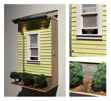 Miniature model of a yellow house wall with window and bushes, showing a hidden detail behind the window.