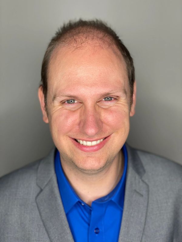 Headshot of James looking at camera an smiling. He is wearing a gray suit and blue shirt.