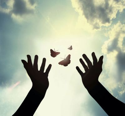 Silhouetted hands reaching towards butterflies in the bright sky.