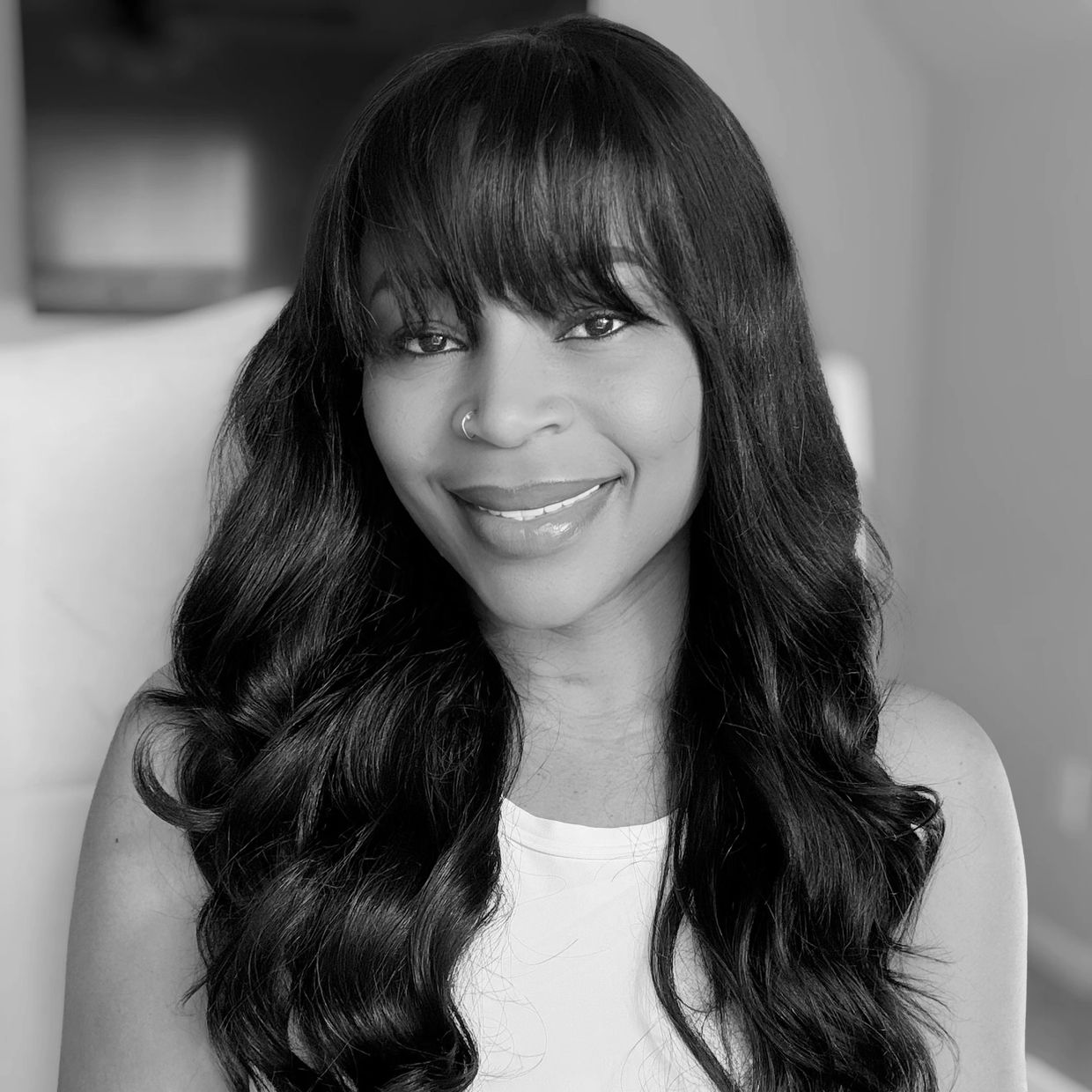 Smiling woman with long wavy black hair and bangs, wearing a white sleeveless top.