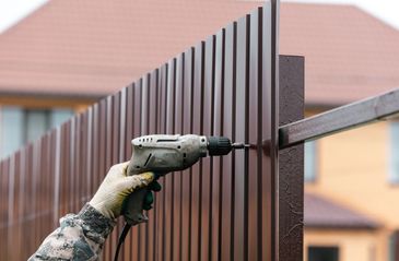 Hand in work gloves using a drill to install a brown metal fence