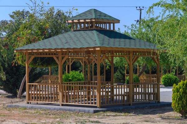 A sturdy wooden gazebo with a dark green shingled roof surrounded by green foliage under a clear sky