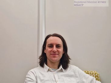 Man with long hair in a white shirt sitting on a patterned chair.