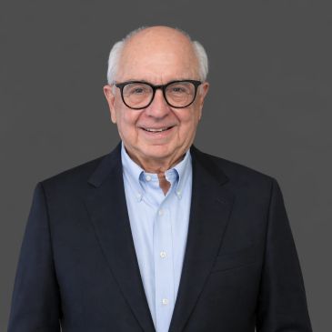 Smiling elderly man in glasses wearing a suit jacket and light blue shirt.