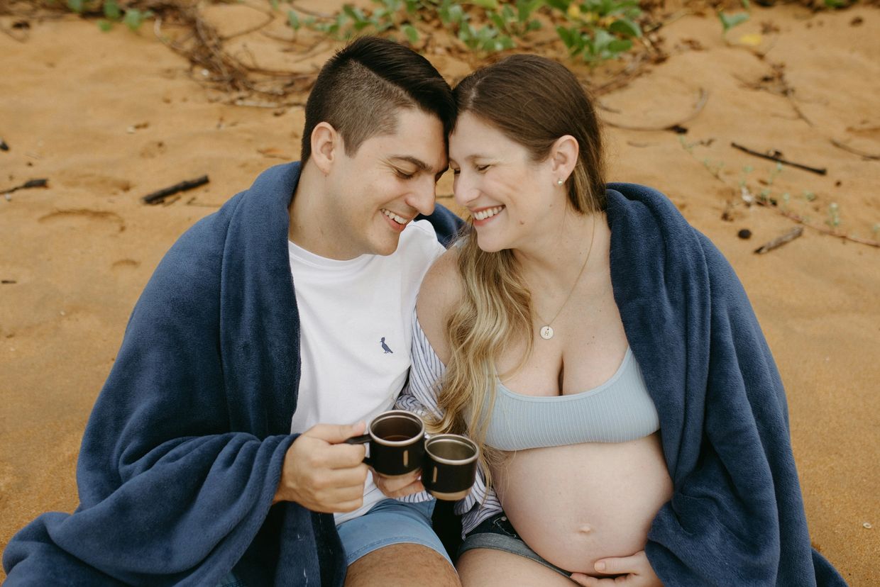 A couple cuddled up in a blanket on the beach, the woman is pregnant