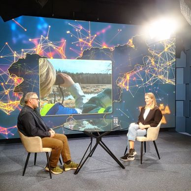 Two people sit in a studio with a large screen behind them showing a nature scene.