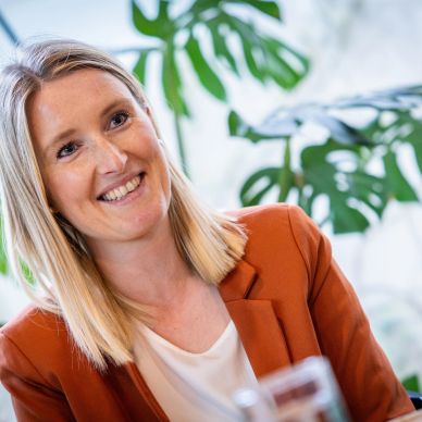 Smiling woman in an orange blazer in a bright room with plants.