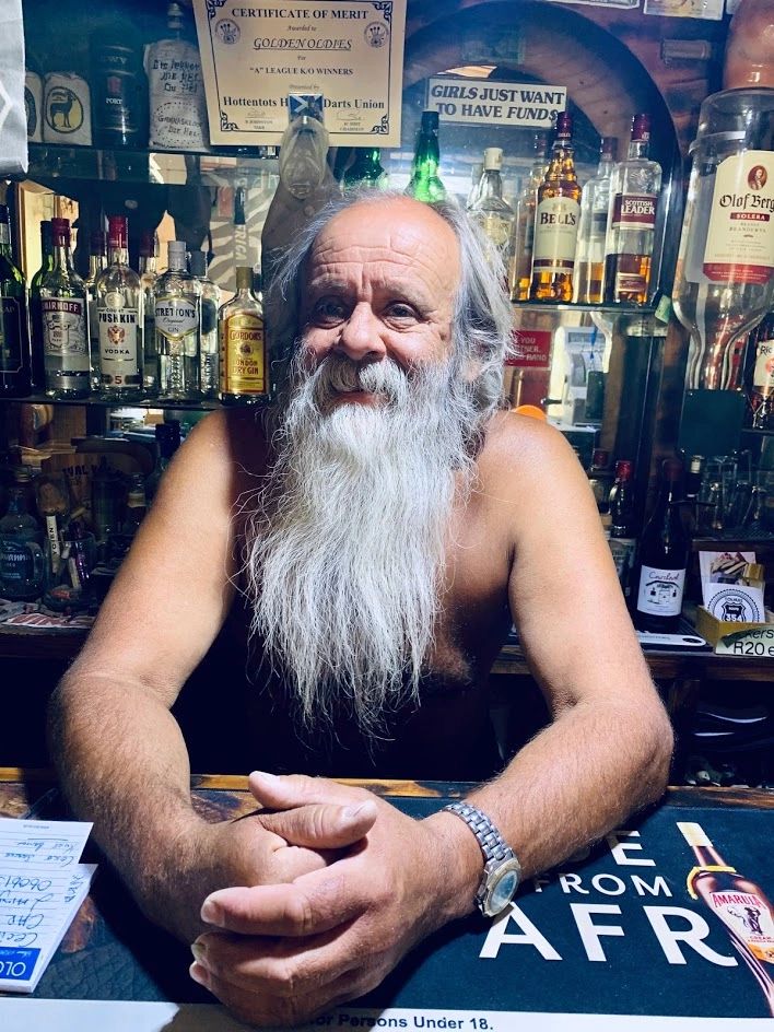 Elderly man with long white beard behind a bar counter, surrounded by bottles and signs.