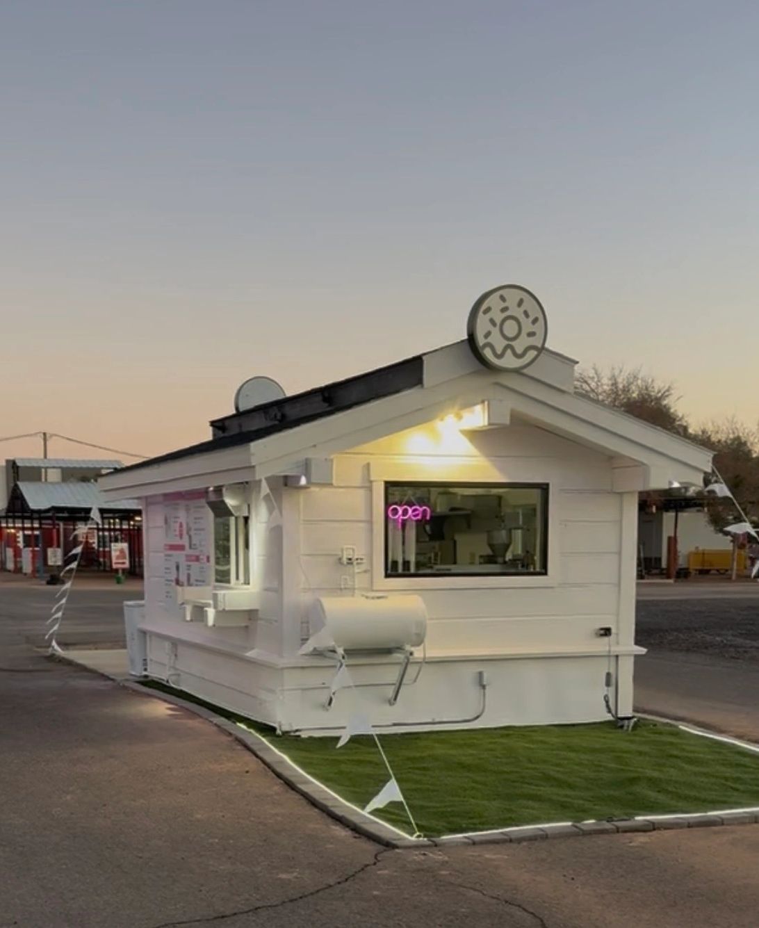 Small white donut shop with neon open sign at dusk.