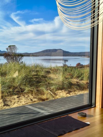 View of a serene lake and mountain through a large window with a wooden deck and grass.